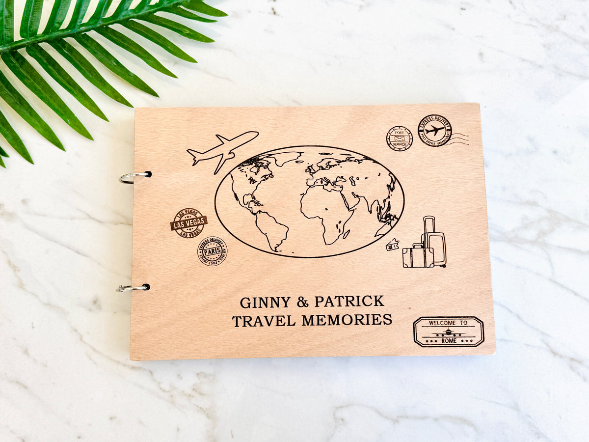 A personalized wooden travel memory book with engraved globe, passport stamp, suitcase, and airplane illustrations, titled 'GINNY & PATRICK TRAVEL MEMORIES,' photographed on a white marble surface with a green palm leaf in the corner.