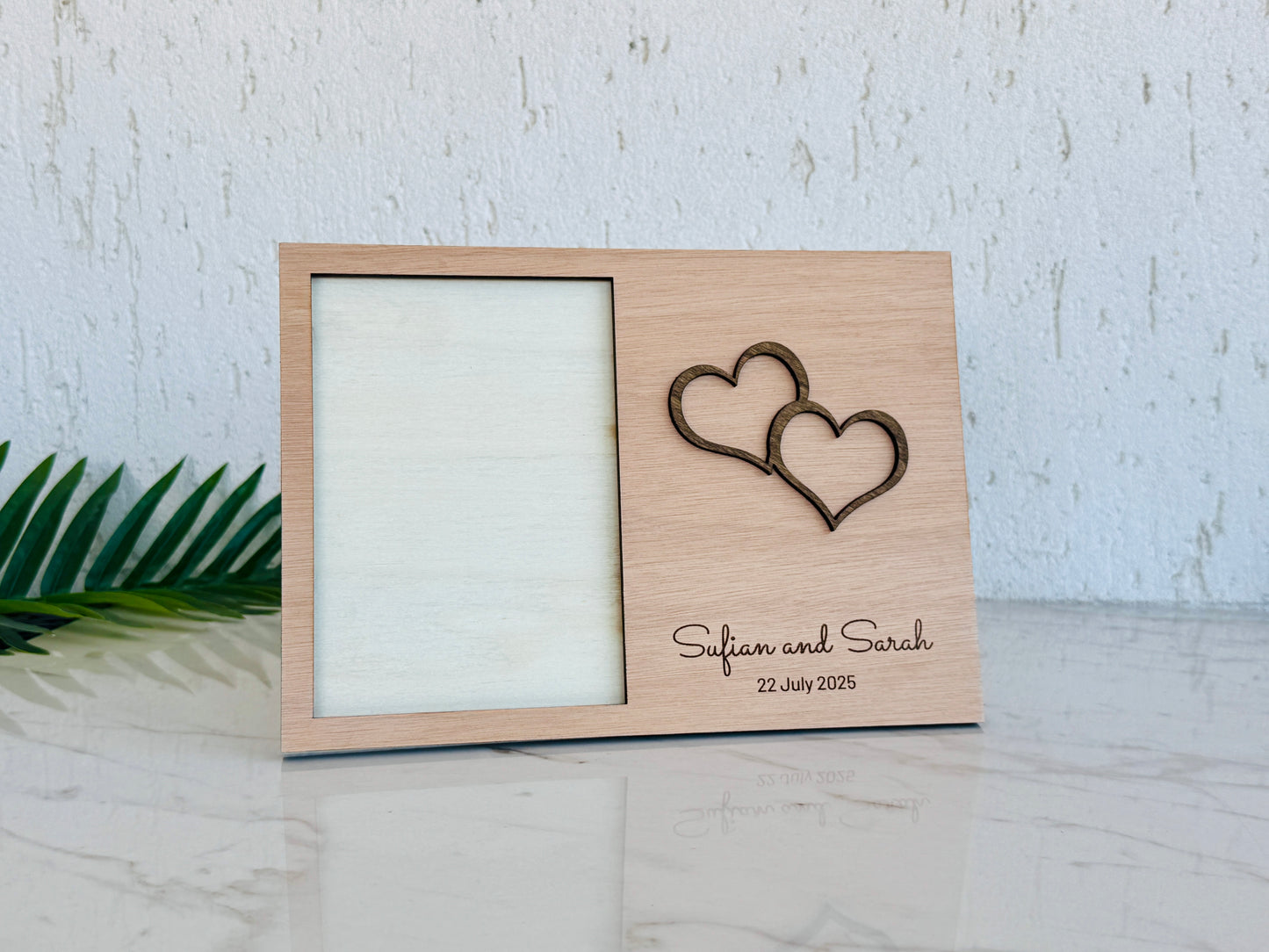 A personalized light wood picture frame with a laser-engraved double heart design and the names 'Safian and Sarah' with the date '22 July 2025' on the front, displayed on a marble surface against a textured white wall.