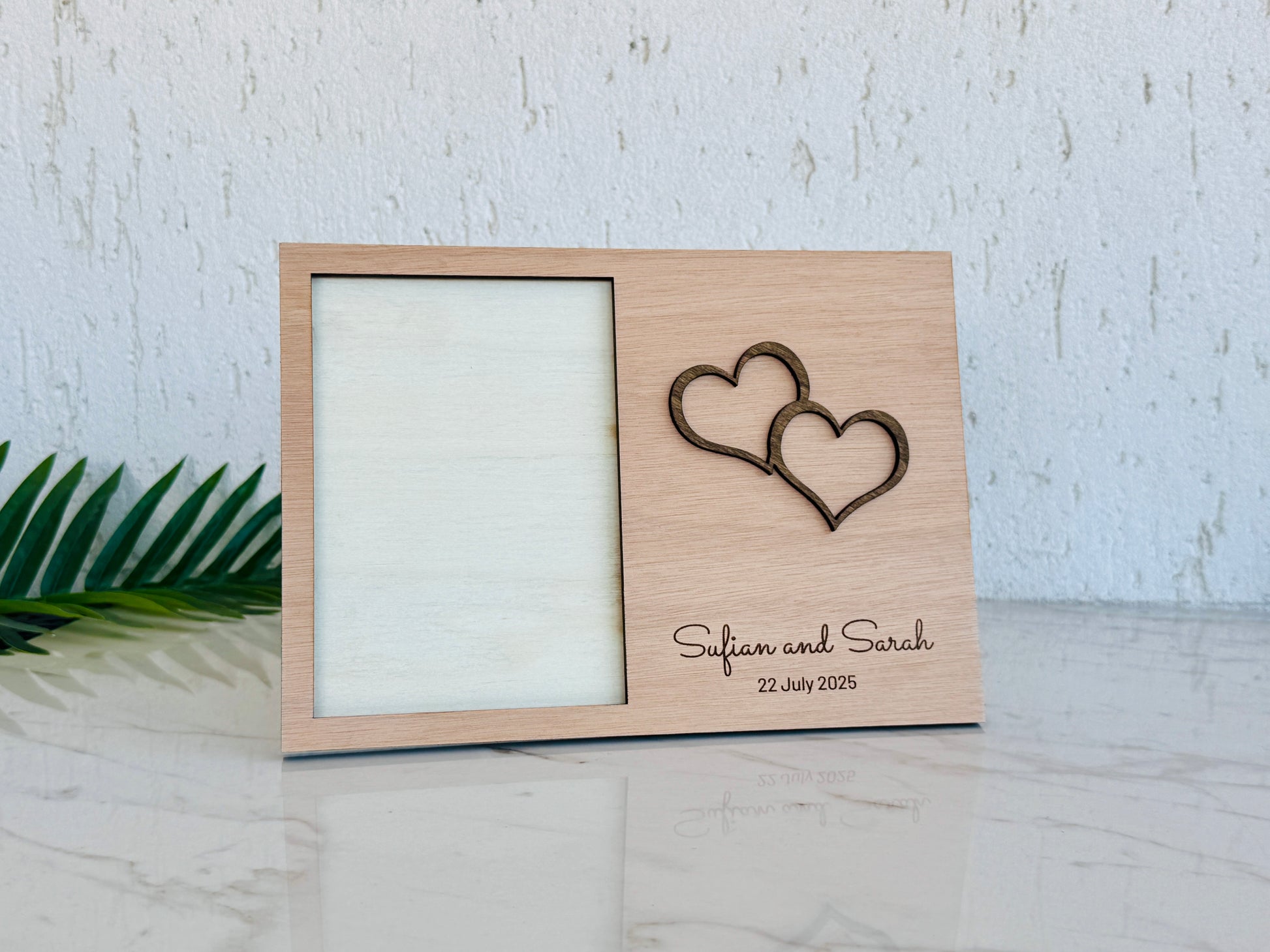 A personalized light wood picture frame with a laser-engraved double heart design and the names 'Safian and Sarah' with the date '22 July 2025' on the front, displayed on a marble surface against a textured white wall.