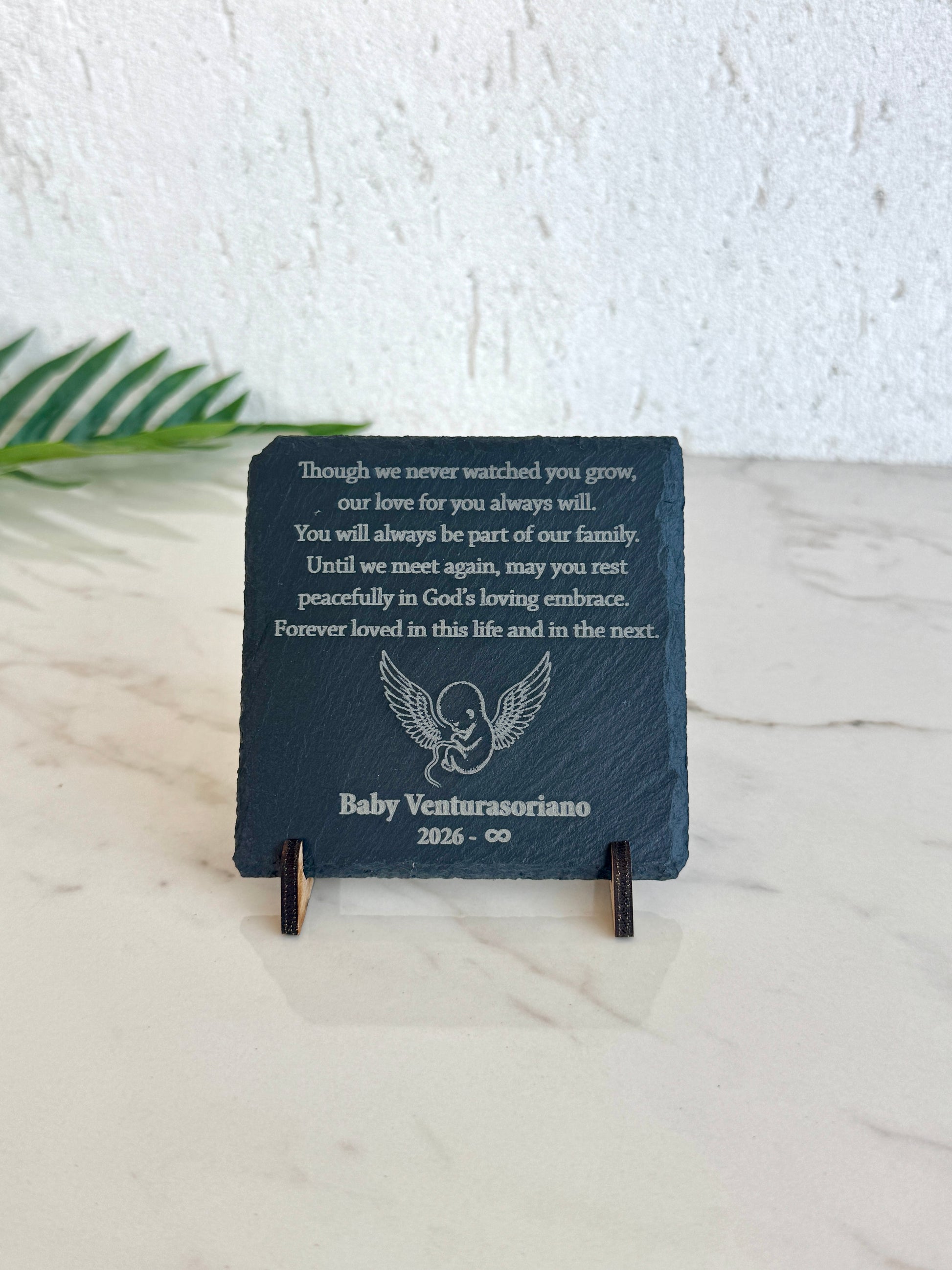 A dark slate memorial plaque on a small wooden display stand, featuring an engraved remembrance poem for Baby Venturasio and a small angel illustration.