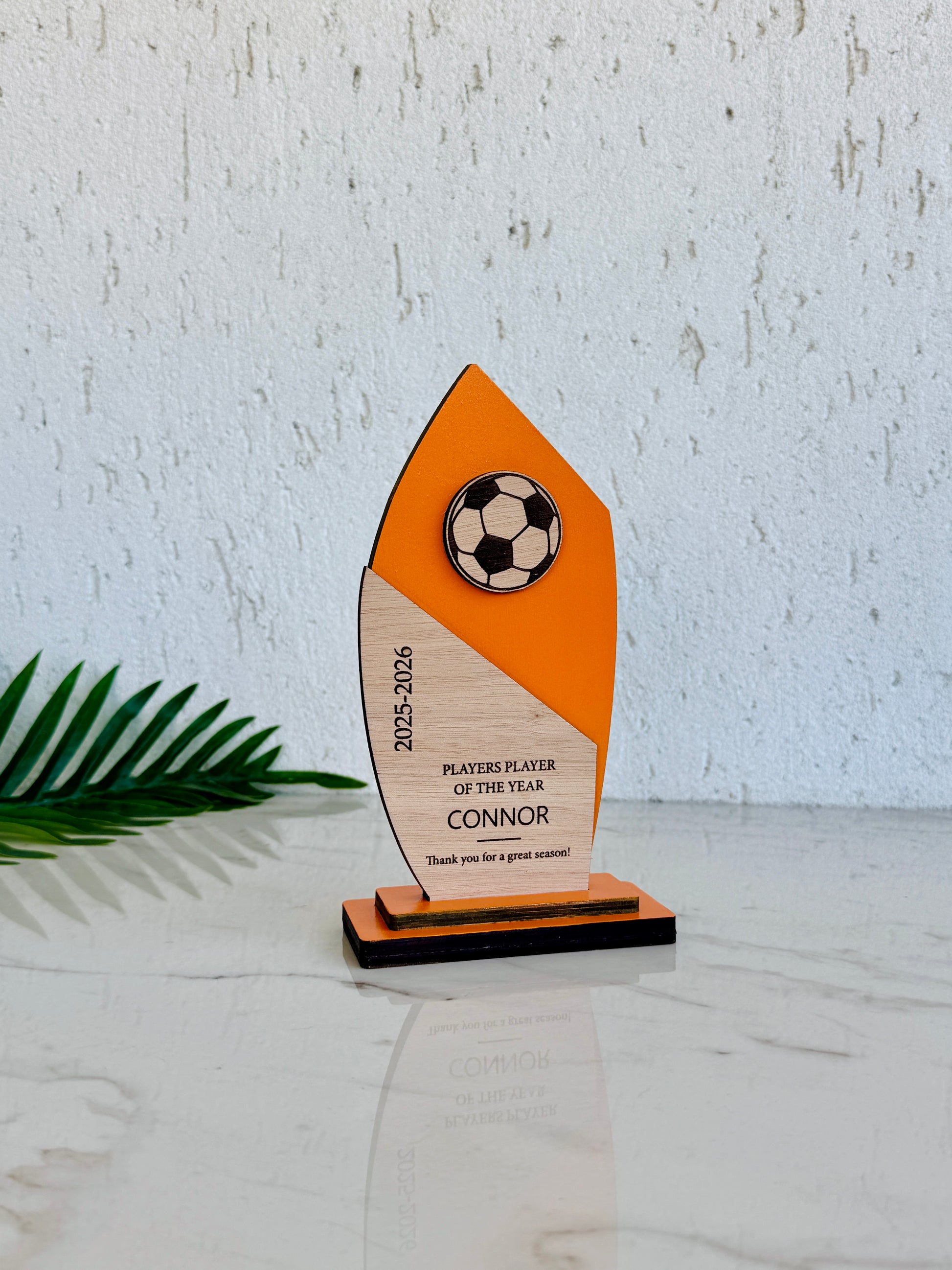 A custom wooden soccer player trophy with an orange and natural wood design, featuring a soccer ball graphic and personalized text, displayed on a marble surface against a textured white wall.