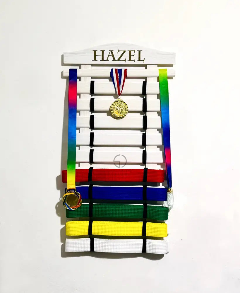 Personalized wooden martial arts belt display rack with engraved name ‘Lincoln’, in white color holding karate or taekwondo belts along with hanging medals on top slat. Perfect wall-mounted martial arts belt holder for kids’ achievements and sports awards organization. Available in various colors and sizes