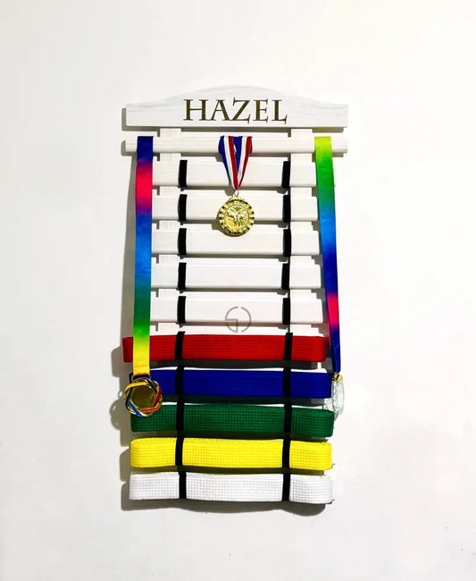 Personalized wooden martial arts belt display rack with engraved name ‘Lincoln’, in white color holding karate or taekwondo belts along with hanging medals on top slat. Perfect wall-mounted martial arts belt holder for kids’ achievements and sports awards organization. Available in various colors and sizes