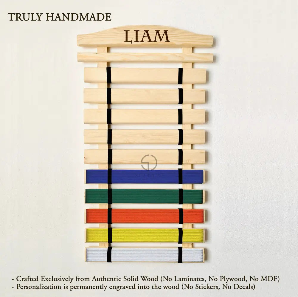 Handmade wooden martial arts belt display rack mounted on a wall, featuring multiple horizontal slats holding colored belts in rank order. The top slat is engraved with the name "Liam" and includes space for hanging medals. Made from solid wood, the rack is available in various sizes (5–18 belt slots plus medal slat) and colors including natural wood, Palisander dark brown, Graphite gray, white, gray, pink, and blue. Designed for wall mounting and personalized with laser engraving by Shiandecreations