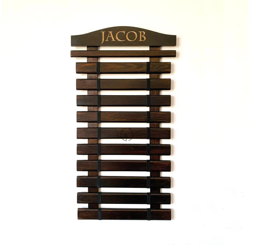 Handmade wooden martial arts belt display rack mounted on a wall, featuring multiple horizontal slats holding colored belts in rank order. The top slat is engraved with the name "Jacob" and includes space for hanging medals. Made from solid wood, the rack is available in various sizes (5–18 belt slots plus medal slat) and colors including natural wood, Palisander dark brown, Graphite gray, white, gray, pink, and blue. Designed for wall mounting and personalized with laser engraving by Shiandecreations