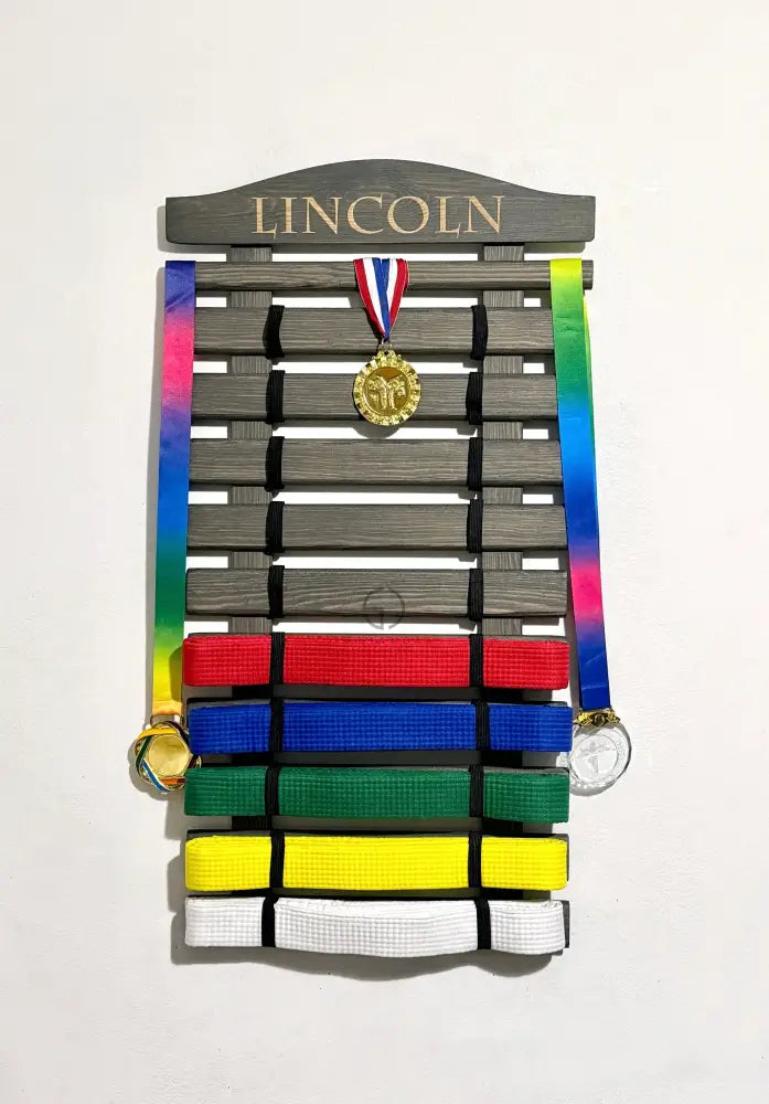 Personalized wooden martial arts belt display rack with engraved name ‘Lincoln’, in graphite gray color holding karate or taekwondo belts along with hanging medals on top slat. Perfect wall-mounted martial arts belt holder for kids’ achievements and sports awards organization. Available in various colors and sizes. Ships worldwide
